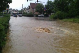 Überflutete Straße ist zu sehen, Wasser fließt in einen Kanal. Hier muss der Hochwasserschutz verbessert werden.